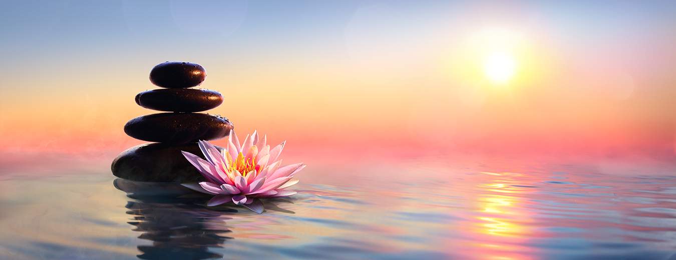 A serene sunset scene over calm water featuring a stack of balanced black stones next to a blooming pink lotus flower