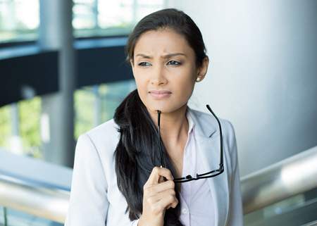 A therapist with a thoughtful expression, holding her glasses in one hand, standing in a modern office environment, providing an Individual Therapy Service