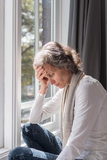 A woman sitting by a window with her head resting in her hand, appearing thoughtful and distressed, with natural light streaming in