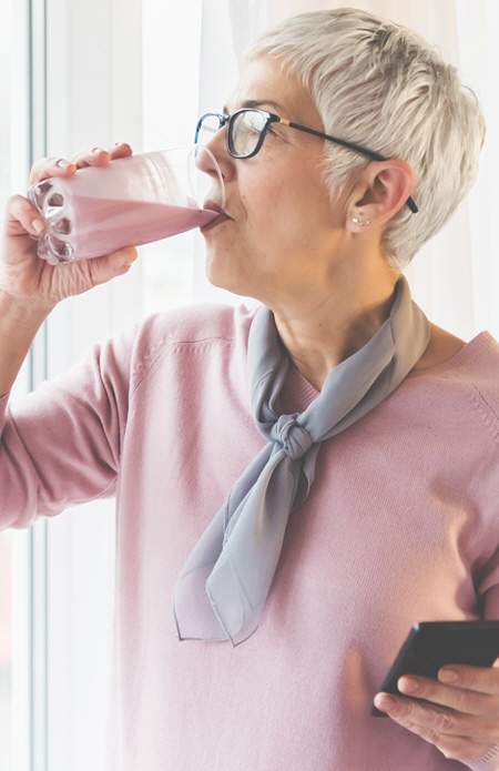 A former client named 'Sally' wearing glasses and a light pink sweater, drinking a smoothie while holding a phone in her other hand