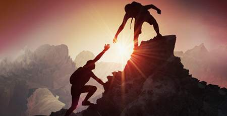 Two climbers silhouetted on a mountain peak at sunset, with one reaching down to help the other up, symbolizing support and teamwork