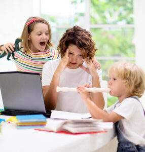 A stressed parent sits at a table with a laptop and paperwork, holding their head, while two young children play noisily around them, symbolizing work-life balance challenges