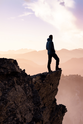 A person standing on the edge of a rocky cliff at sunrise or sunset, overlooking a vast mountainous landscape