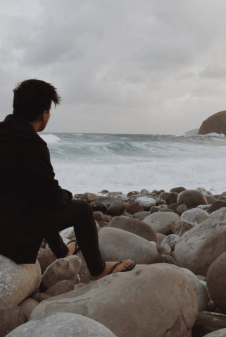 A person sitting on large rocks at the edge of a beach, gazing out at the ocean waves under a cloudy sky