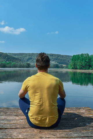 A man in a yellow shirt sitting cross-legged on a wooden dock, gazing out at a calm lake surrounded by trees and hills on a clear day