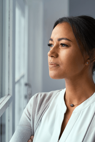 A woman standing with arms crossed, gazing thoughtfully out of a window, wearing a light blouse and a soft cardigan