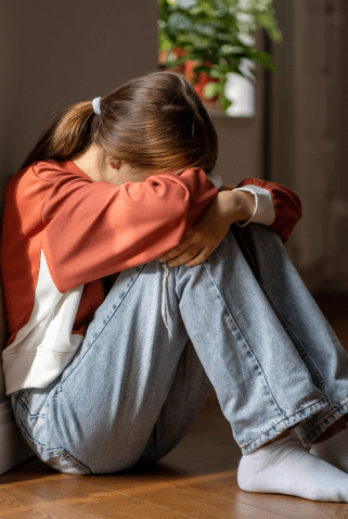 A young person sitting on the floor with their head resting on their knees, arms wrapped around their legs, appearing distressed, wearing a red sweatshirt and jeans