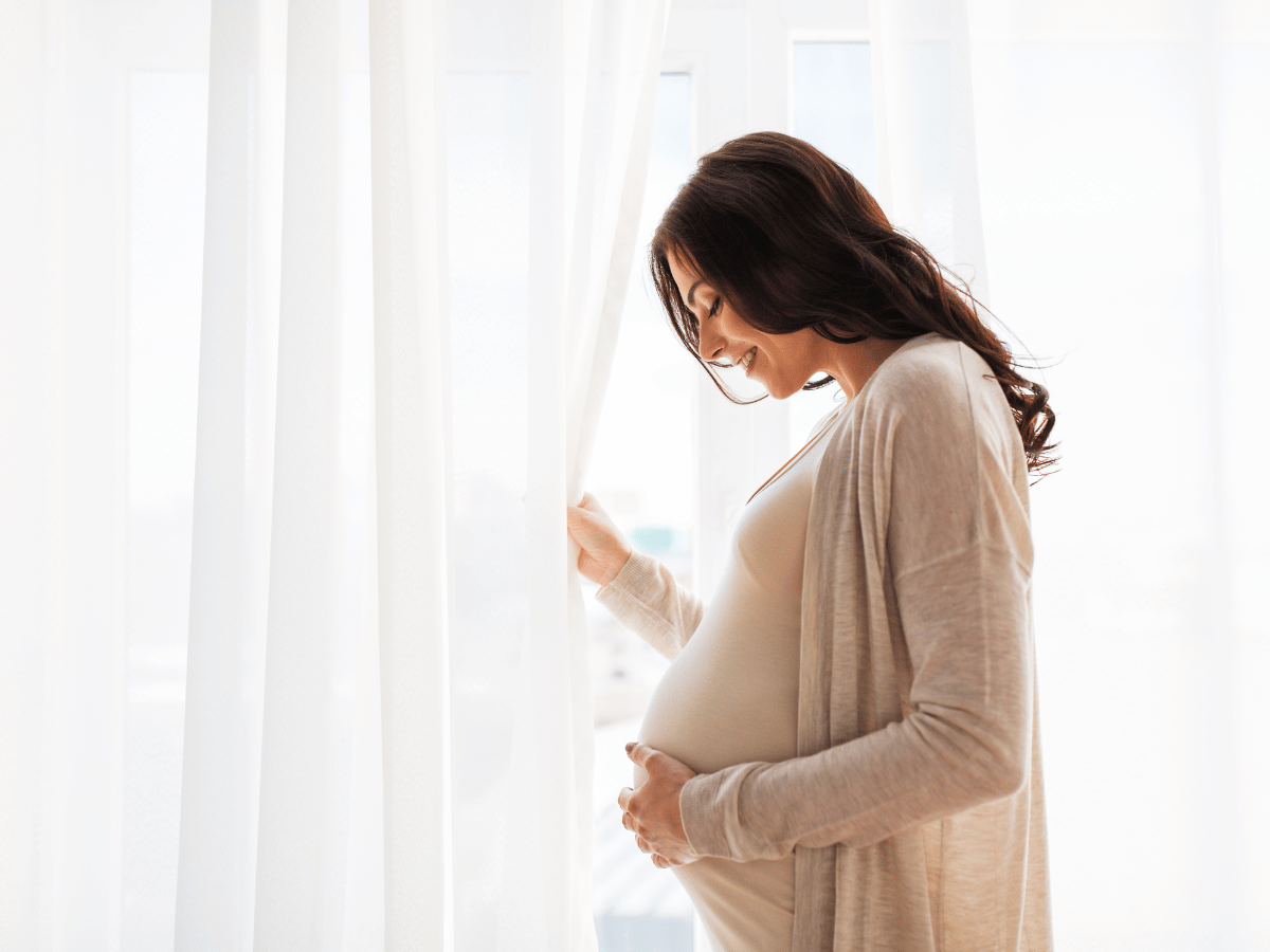 A pregnant woman standing by a window, gently holding her belly and smiling, with soft light coming through white curtains