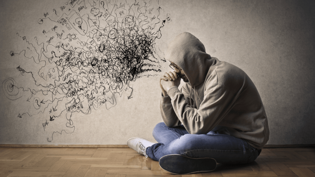 A person sitting with their head in their hands, surrounded by chaotic scribbles representing negative self-talk, mental health struggles, and self-dislike.