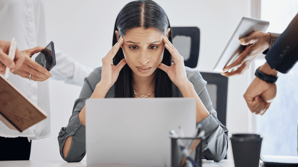 Woman at a desk looking overwhelmed, hands on her temples as multiple people present her with tasks and documents