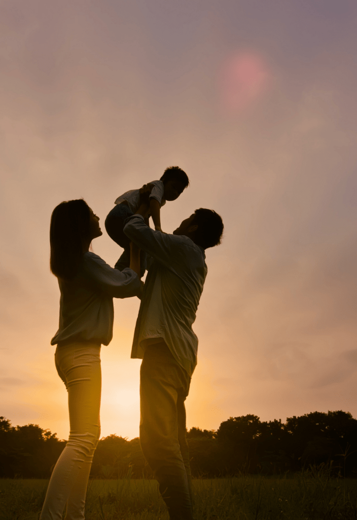 A silhouette of a family of three is captured against a warm, golden sunset in an open field. The father lifts their young child into the air while the mother stands close, supporting them and smiling. The sky has soft hues of orange and pink, with a gentle glow surrounding the scene, evoking a sense of love, connection, and joy.