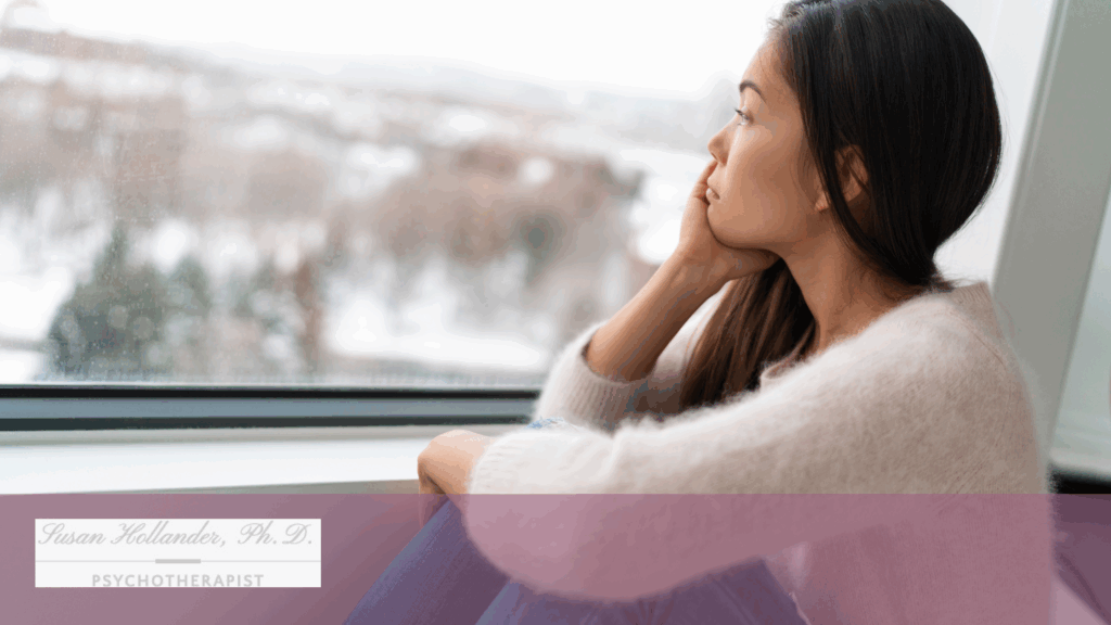 A young woman sits by a window on a winter day, gazing outside with a pensive, distant expression.