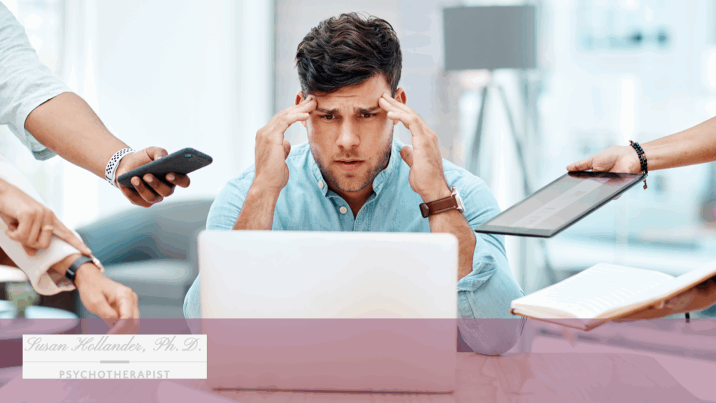 Frustrated man sitting at a laptop with hands on his temples as multiple coworkers demand his attention, symbolizing workplace stress and emotional overload.