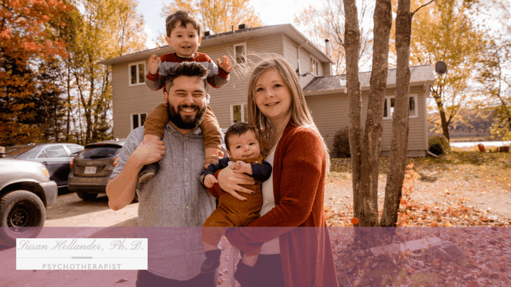 A smiling couple stands outside a home with their two young children, surrounded by autumn trees and fallen leaves.