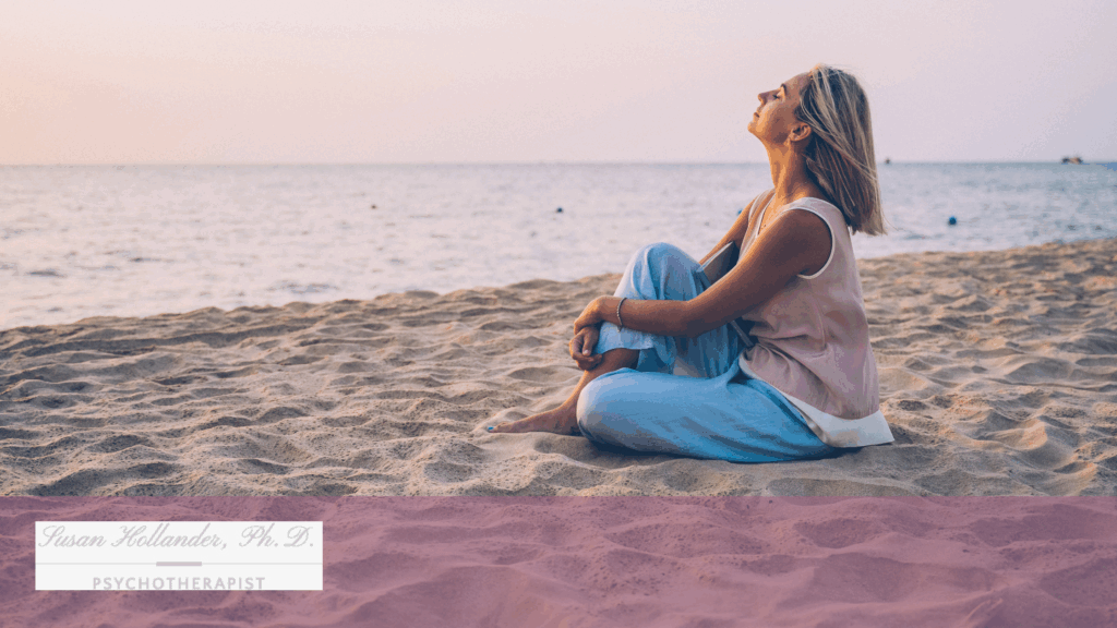 Woman sitting peacefully on the beach at sunset, looking upward with a relaxed expression, symbolizing calm and emotional balance.